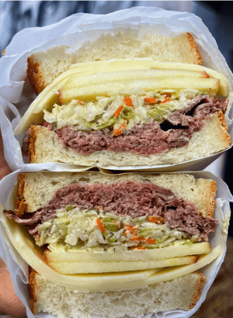 A close-up of a Power City Eatery sandwich cut in half, revealing layers of roast beef, coleslaw with chopped vegetables, slices of white cheese, and thick white bread.